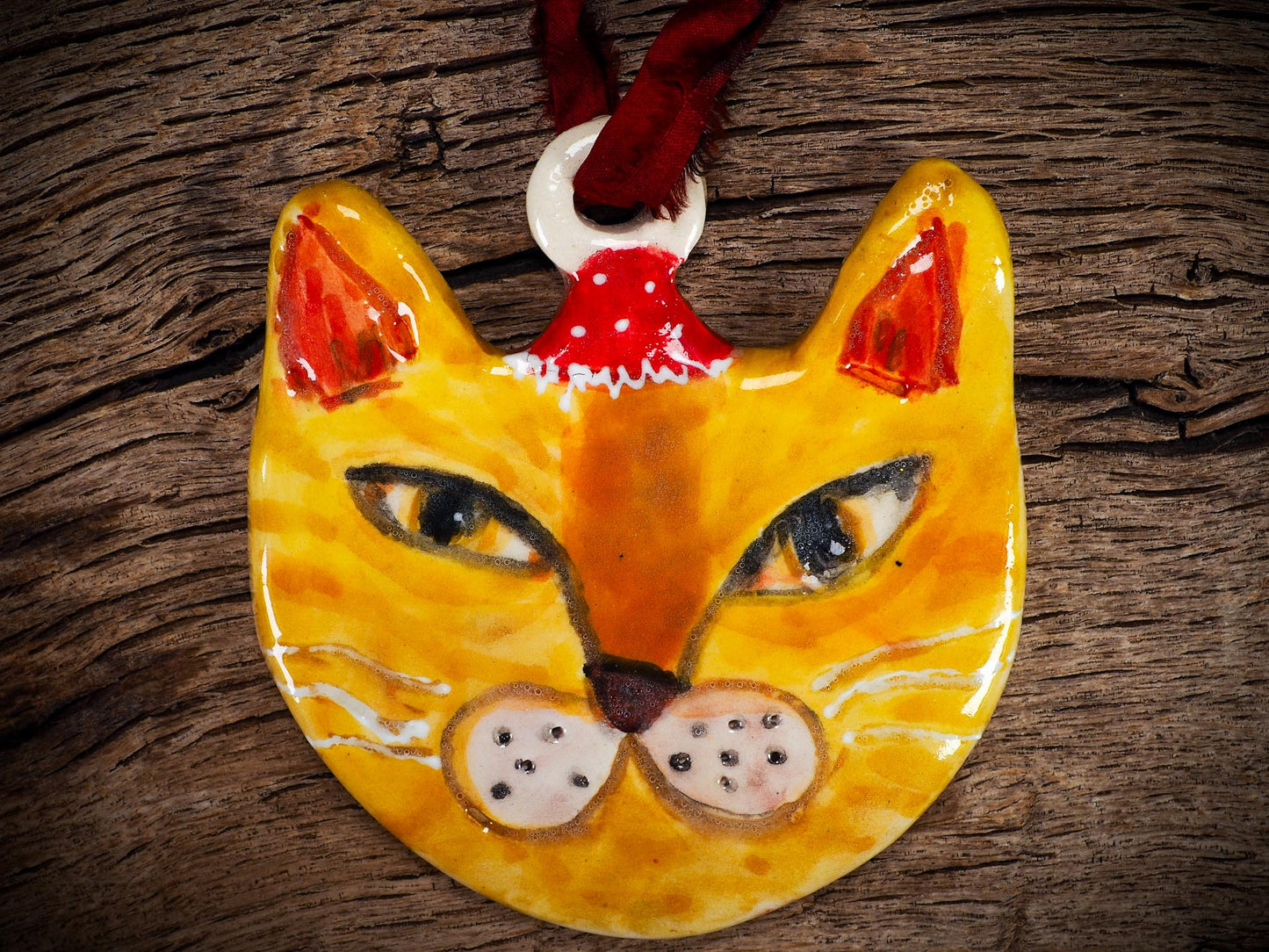 Handmade ceramic ginger cat ornament with red holiday hat and ribbon, created by Idania Salcido Danita Art for Christmas decorating or gifting.
