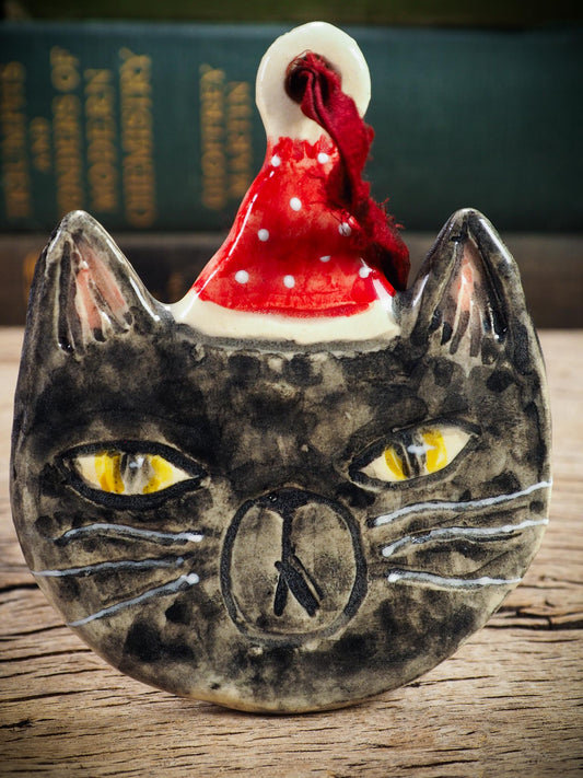 Handmade ceramic black cat ornament named Loki with red holiday hat and ribbon, created by Danita Art Idania Salcido for Christmas decorating or gifting.