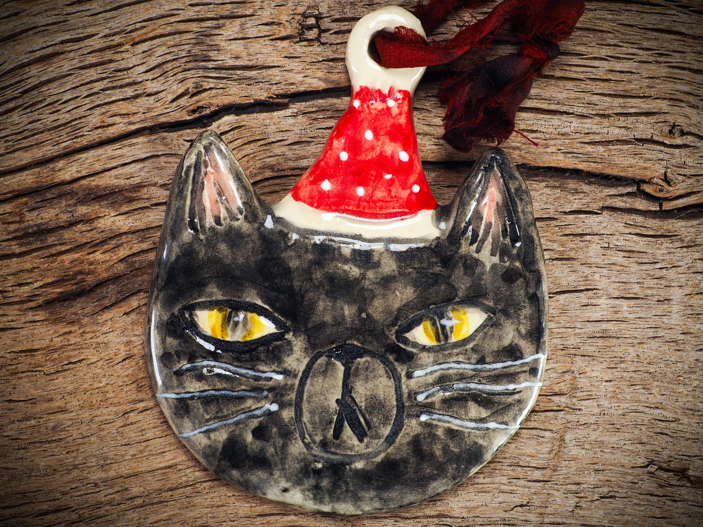 Handmade ceramic black cat ornament named Loki with red holiday hat and ribbon, created by Danita Art Idania Salcido for Christmas decorating or gifting.