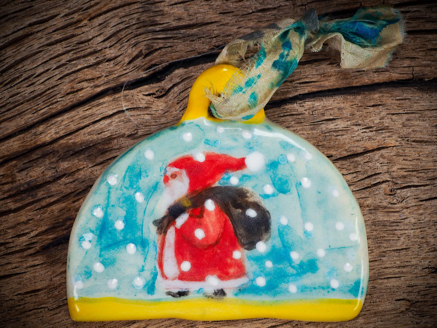Handmade ceramic Santa ornament with snowy scene and ribbon, created by Idania Salcido Danita Art for Christmas decoration or gifting.