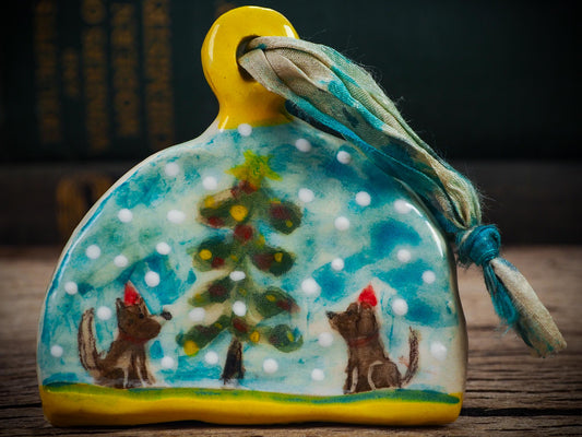 Handmade ceramic winter dogs ornament with snowy scene and ribbon, created by Idania Salcido Danita Art for Christmas decorating or gifting.