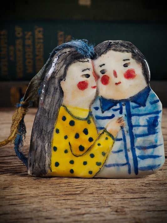 Handmade ceramic winter couple ornament with painted scarves and ribbon, created by Idania Salcido aka Danita Art for Christmas decoration or gifting.