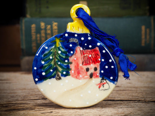 Handmade ceramic winter cottage ornament with snowman and tree, created by Idania Salcido aka Danita Art for Christmas decorating or gifting.
