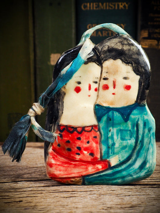 Handmade ceramic winter couple ornament with painted scarves and ribbon, created by Idania Salcido aka Danita Art for Christmas decoration or gifting.