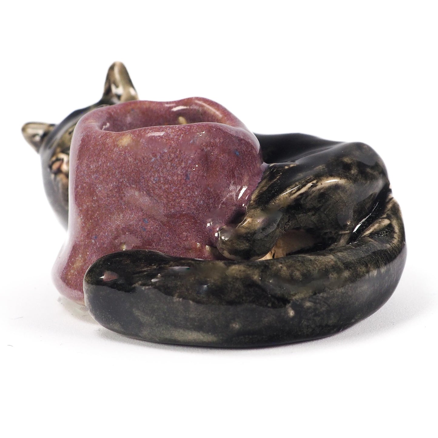 A ceramic cat figurine lying down, holding a small purple bowl or cup in its paws. The cat's head and body are glazed in a dark metallic bronze-black, while the bowl has a textured, speckled purple glaze, by Idania Salcido (Danita Art).