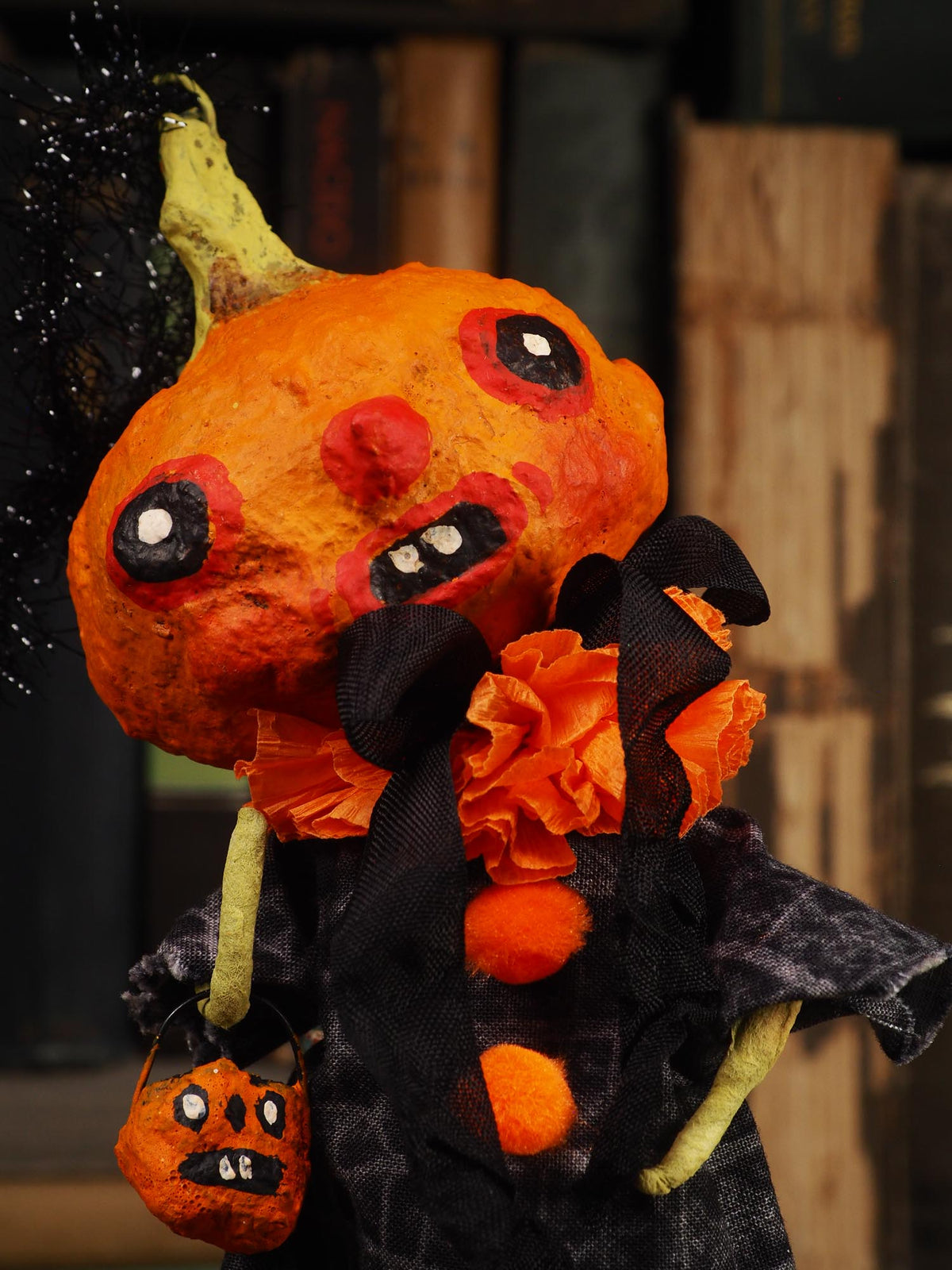 A handmade Halloween pumpkin decoration with a distressed orange body, black and red painted facial features, a green stem, black fabric dress, orange paper flowers, and a small orange pumpkin bucket bag.
An original spooky cute outsider folk art Halloween Doll. Hand made using spun cotton, paper and fabric to create the perfect Halloween ornament for your tree. Specially created for someone who appreciates fine and unique art gifts, It is a one of a kind original design by Idania Salcido (Danita Art).