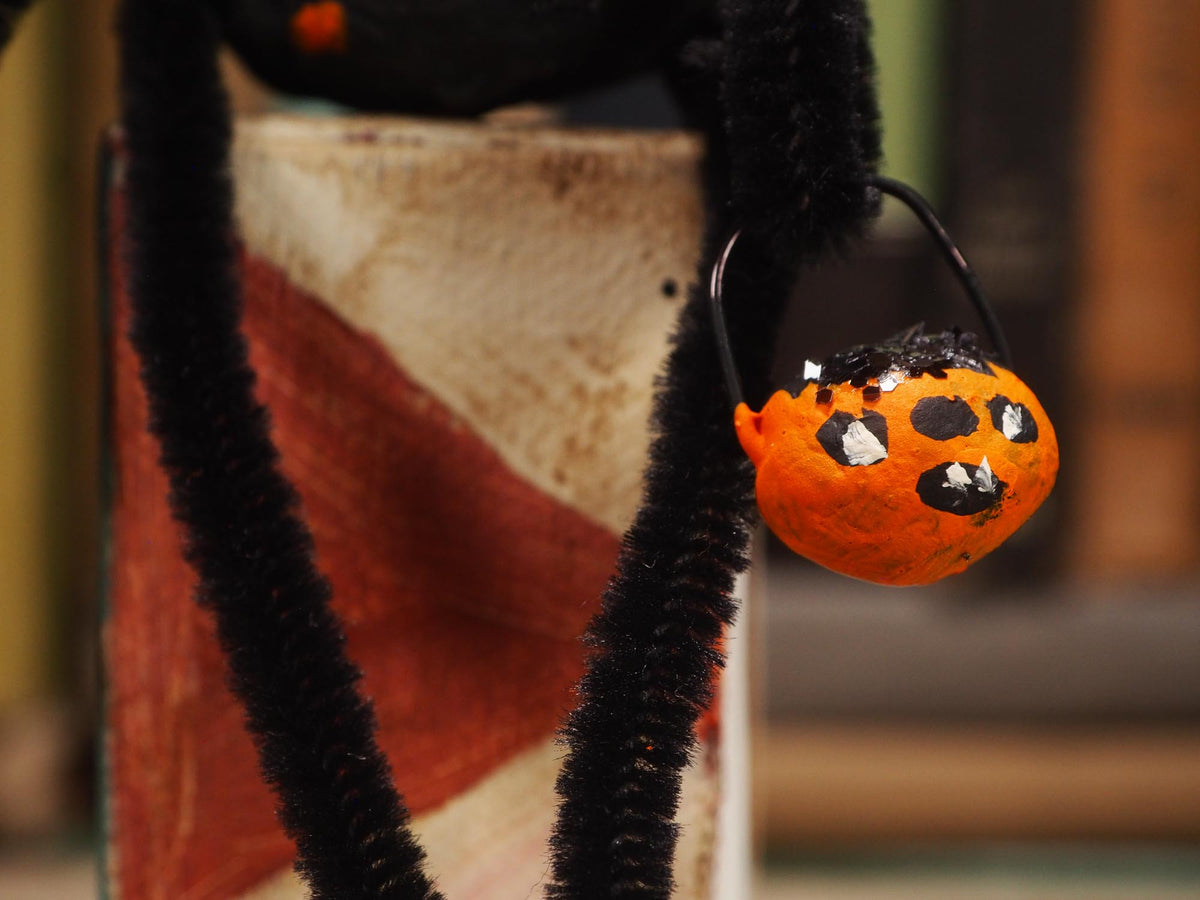 A close-up of a Halloween spider decoration with black fuzzy legs and a small orange spiderweb basket hanging from a black ring. Original spun cotton hand made Halloween doll ornament by Idania Salcido (Danita Art)