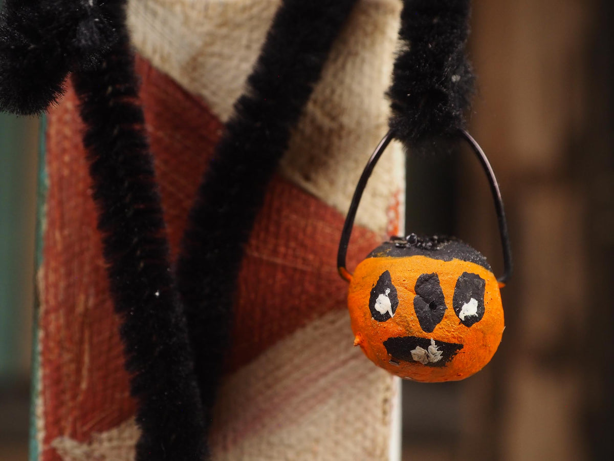 A close-up of a Halloween decoration featuring a small orange pumpkin bucket with a black handle, black eyes, and a black mouth, hanging from a black fuzzy stem against a rustic textile background. Original spun cotton hand made Halloween doll by Idania Salcido (Danita Art)