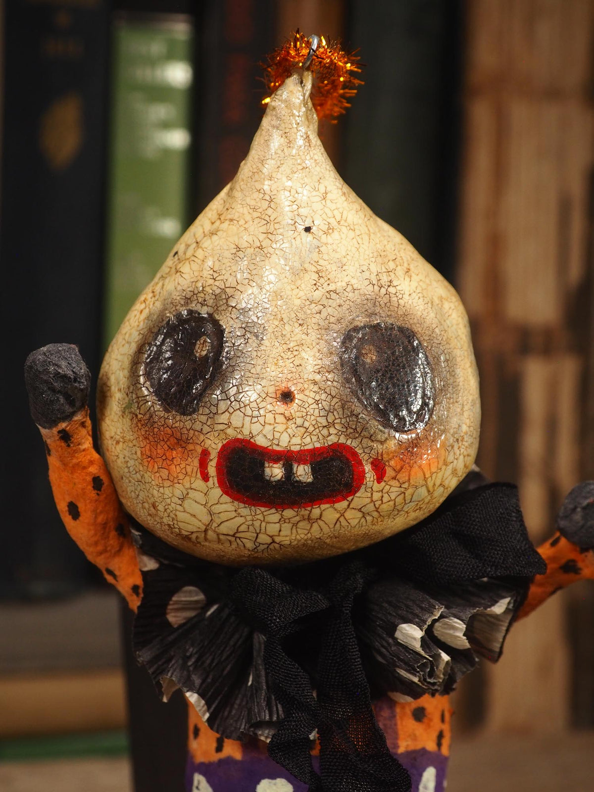 A handmade folk art doll shaped like a cracked beige gourd head with black eyes and a red mouth, wearing an orange and black polka-dot top and purple pants with white dots, standing on a wooden surface with a blurred bookshelf background. An original hand made Halloween art doll by Idania Salcido AKA Danita Art.