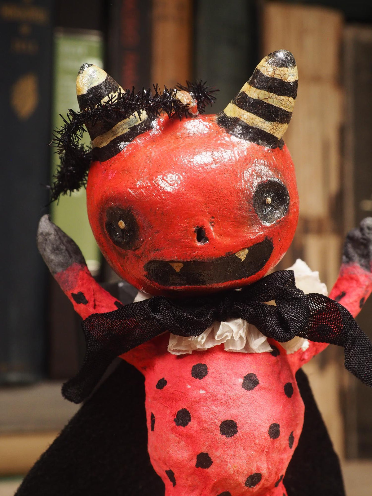 A handmade red and black bat Halloween devil doll with black horns, a black cape, and a ruffled white collar, standing on a wooden surface with a blurred background of books. Original hand made Spun cotton Art Doll by Idania Salcido AKA Danita Art.