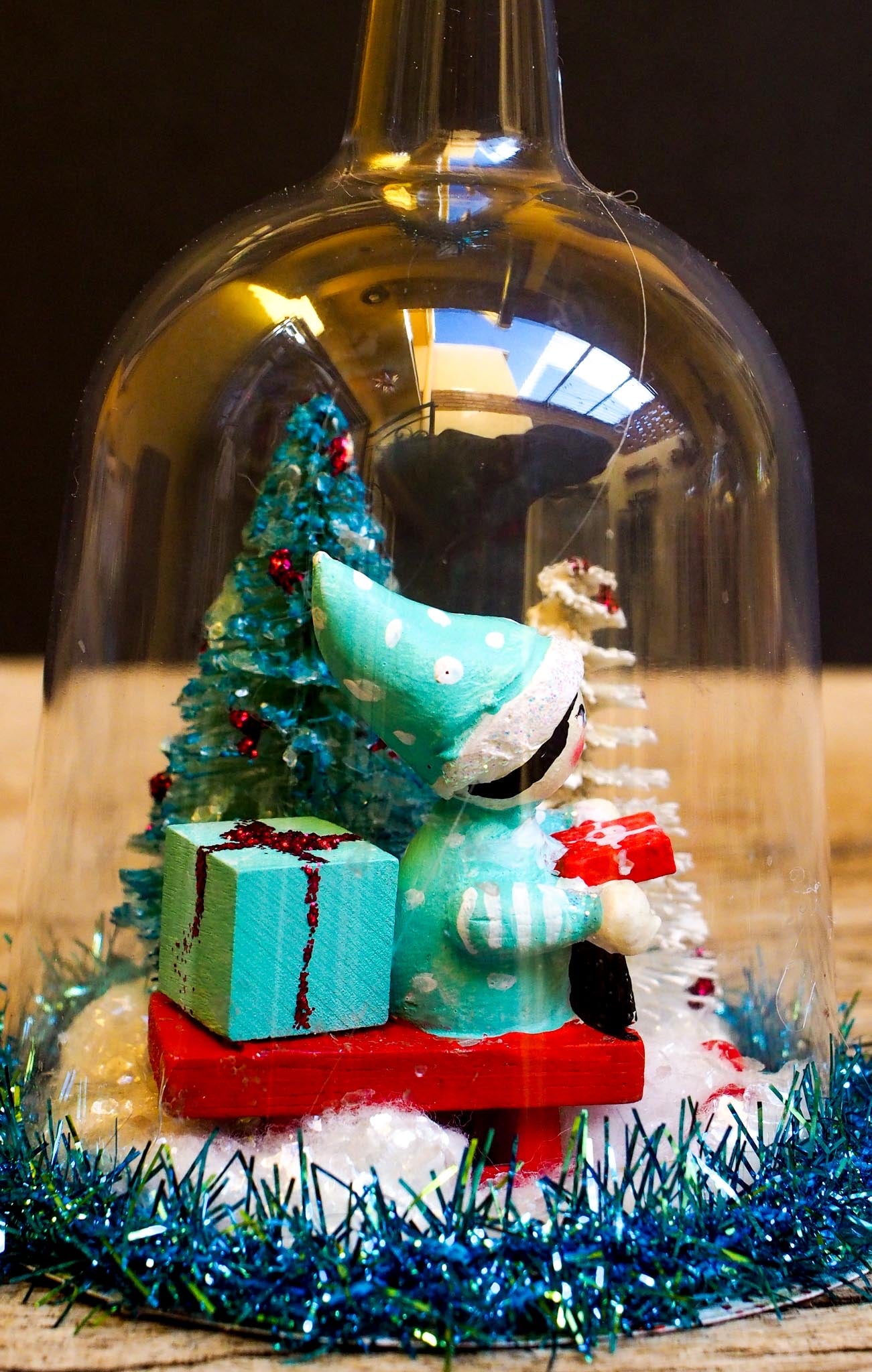 An original handmade christmas tree ornament by Idania Salcido. A clear domed diorama with a holiday scene with Santa Claus elves, Christmas Trees, Elves, Gifts and magical glitter deer.