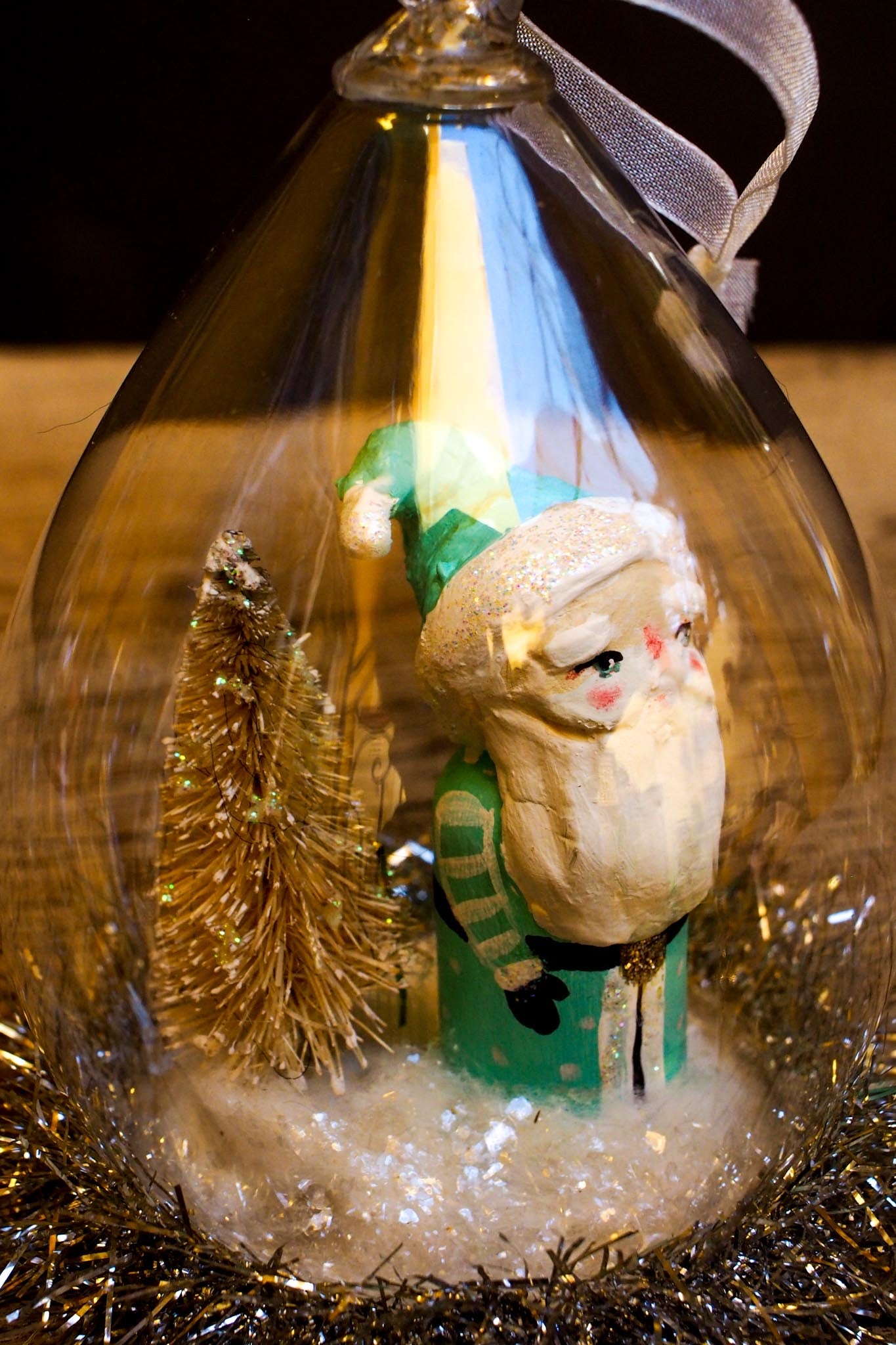 An original handmade christmas tree ornament by Idania Salcido. A clear domed diorama with a holiday scene with Santa Claus elves, Christmas Trees, Elves, Gifts and magical glitter deer.