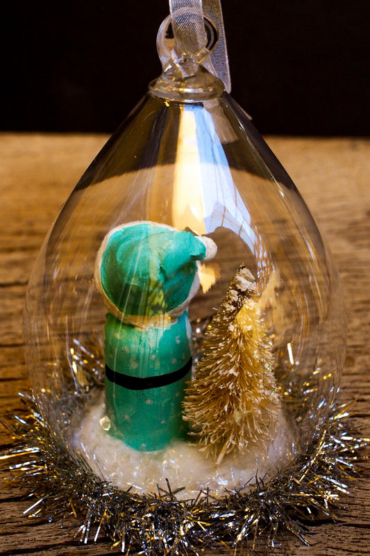 An original handmade christmas tree ornament by Idania Salcido. A clear domed diorama with a holiday scene with Santa Claus elves, Christmas Trees, Elves, Gifts and magical glitter deer.