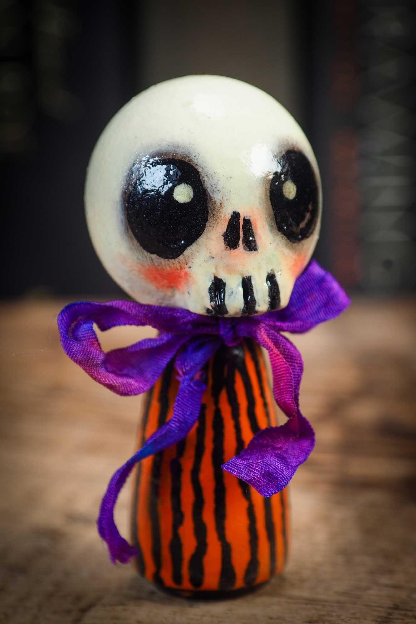 Small Halloween mini kokeshi art doll figurine with skull face, orange and black striped body, and purple ribbon. Made by Idania Salcido, Danita Art.