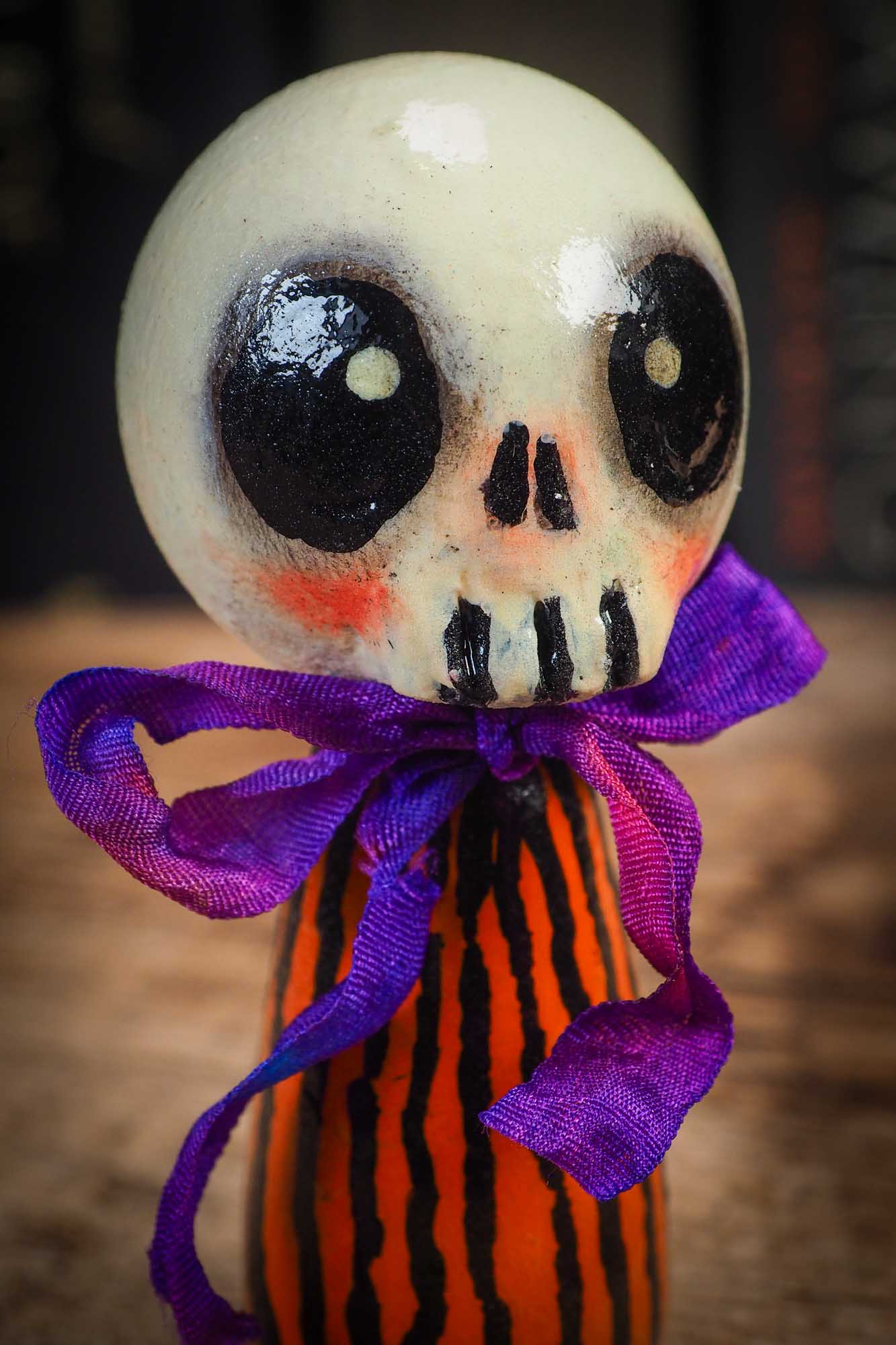 Small Halloween mini kokeshi art doll figurine with skull face, orange and black striped body, and purple ribbon. Made by Idania Salcido, Danita Art.