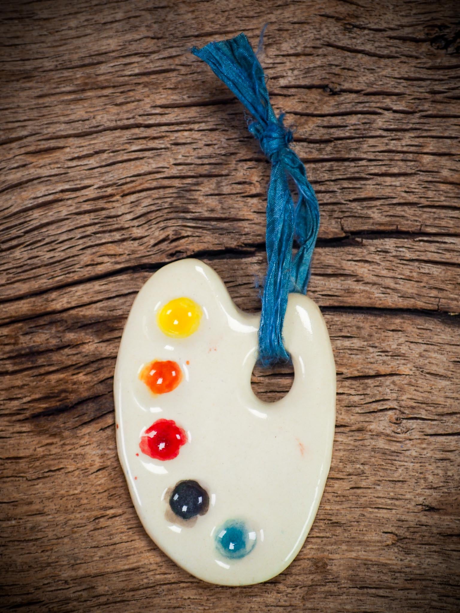 Handmade ceramic mini painter’s palette ornament with colorful paint wells, ideal as an artist gift or Christmas decoration by Idania Salcido Danita Art.
