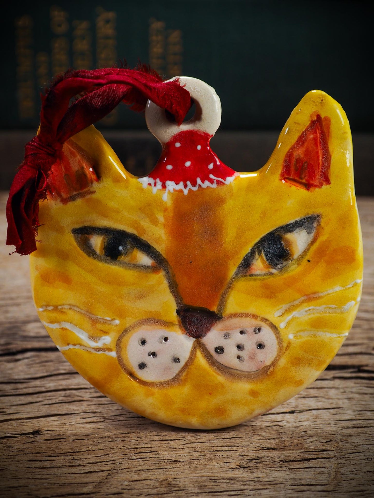 Handmade ceramic ginger cat ornament with red holiday hat and ribbon, created by Idania Salcido Danita Art for Christmas decorating or gifting.
