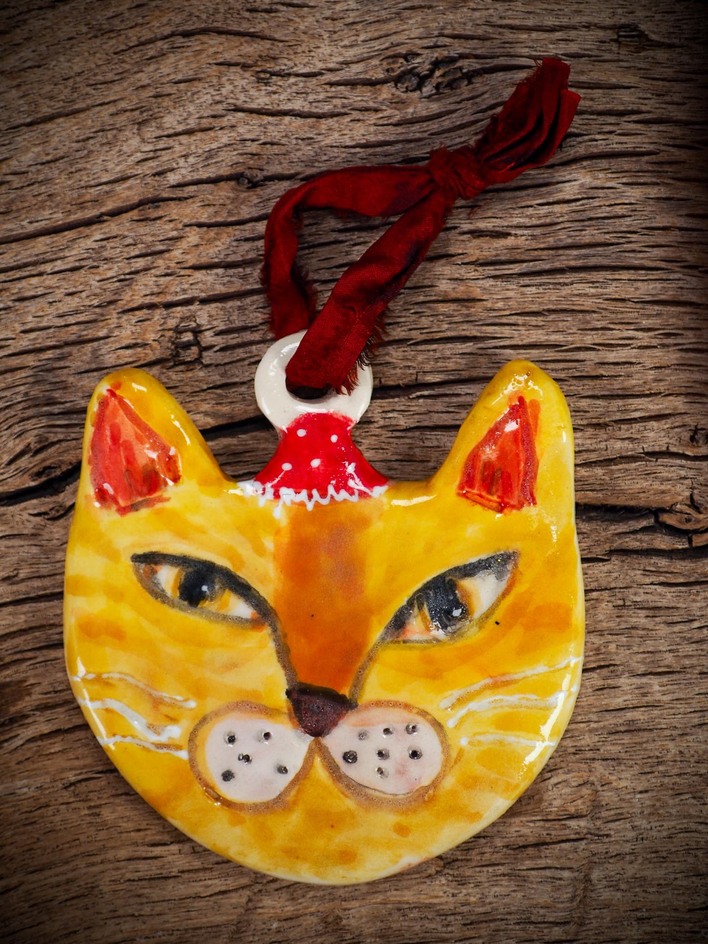 Handmade ceramic ginger cat ornament with red holiday hat and ribbon, created by Idania Salcido Danita Art for Christmas decorating or gifting.