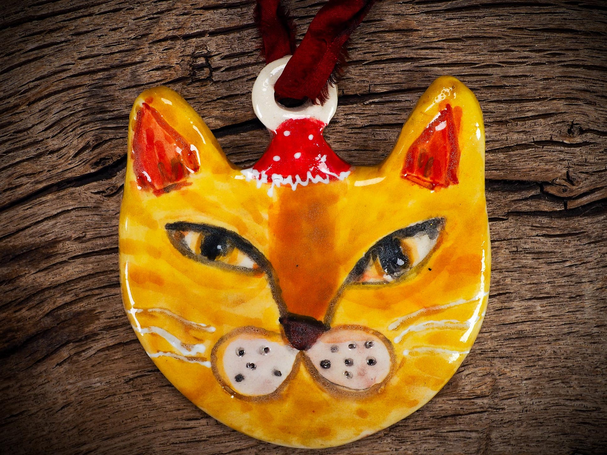 Handmade ceramic ginger cat ornament with red holiday hat and ribbon, created by Idania Salcido Danita Art for Christmas decorating or gifting.