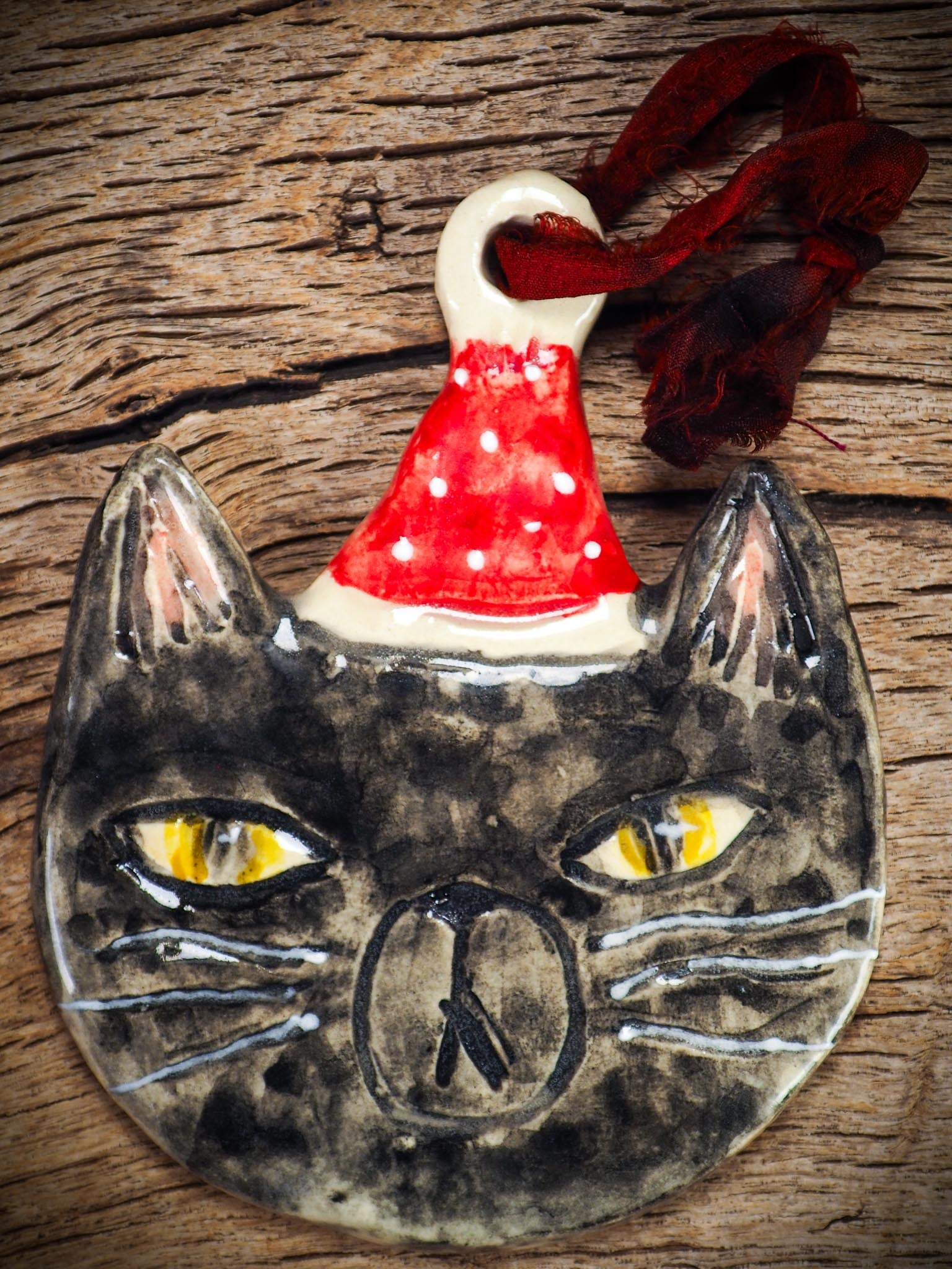 Handmade ceramic black cat ornament named Loki with red holiday hat and ribbon, created by Danita Art Idania Salcido for Christmas decorating or gifting.