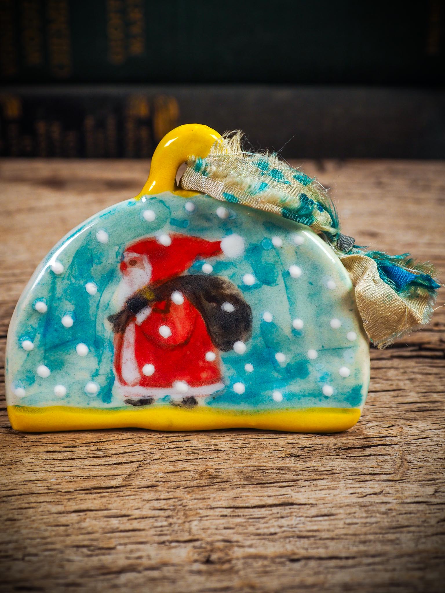 Handmade ceramic Santa ornament with snowy scene and ribbon, created by Idania Salcido Danita Art for Christmas decoration or gifting.