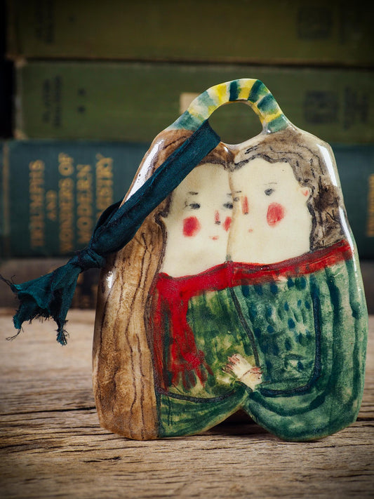 Handmade ceramic winter couple ornament with painted scarves and ribbon, created by Idania Salcido aka Danita Art for Christmas decoration or gifting.