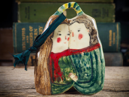 Handmade ceramic winter couple ornament with painted scarves and ribbon, created by Idania Salcido aka Danita Art for Christmas decoration or gifting.