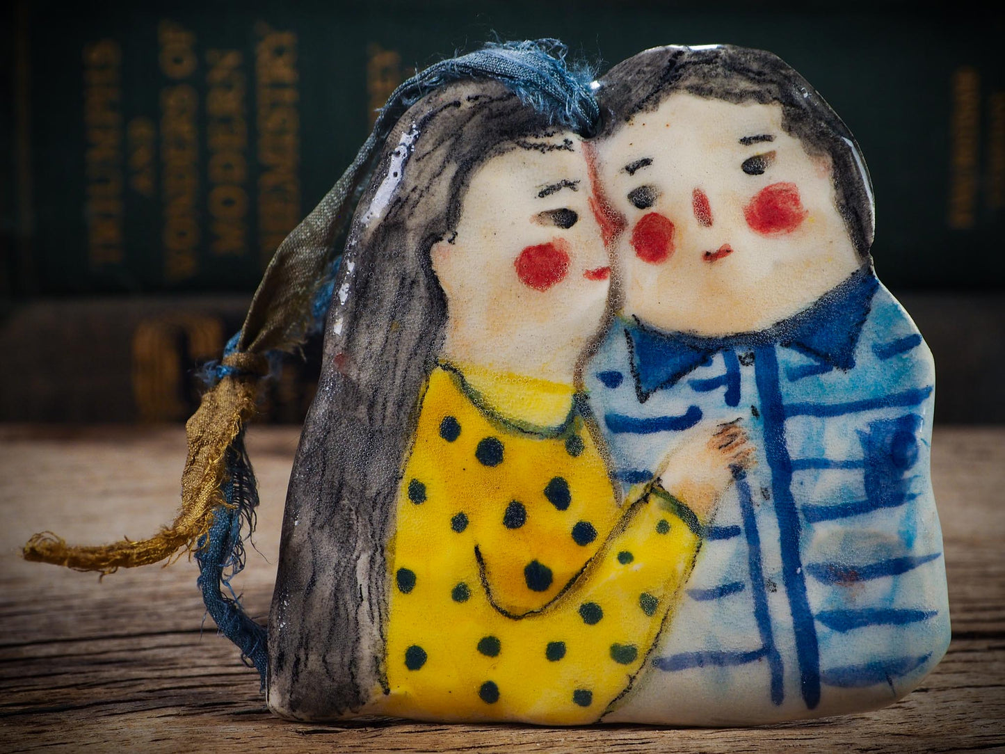 Handmade ceramic winter couple ornament with painted scarves and ribbon, created by Idania Salcido aka Danita Art for Christmas decoration or gifting.