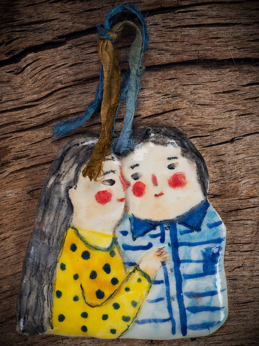 Handmade ceramic winter couple ornament with painted scarves and ribbon, created by Idania Salcido aka Danita Art for Christmas decoration or gifting.