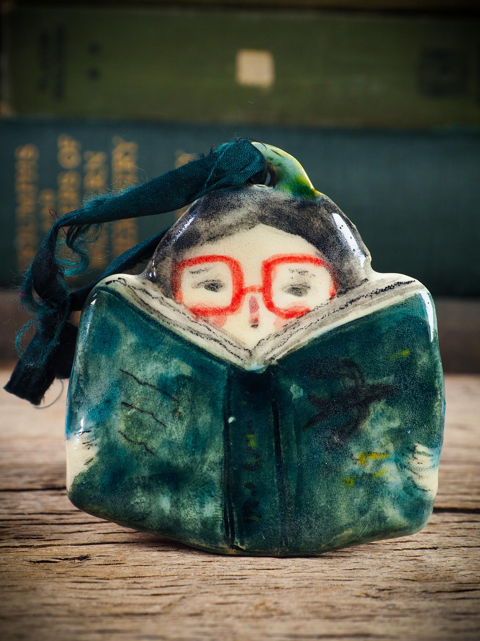 Handmade ceramic reader ornament with red glasses and green book, created by Idania Salcido aka Danita Art for Christmas decorating or gifting.