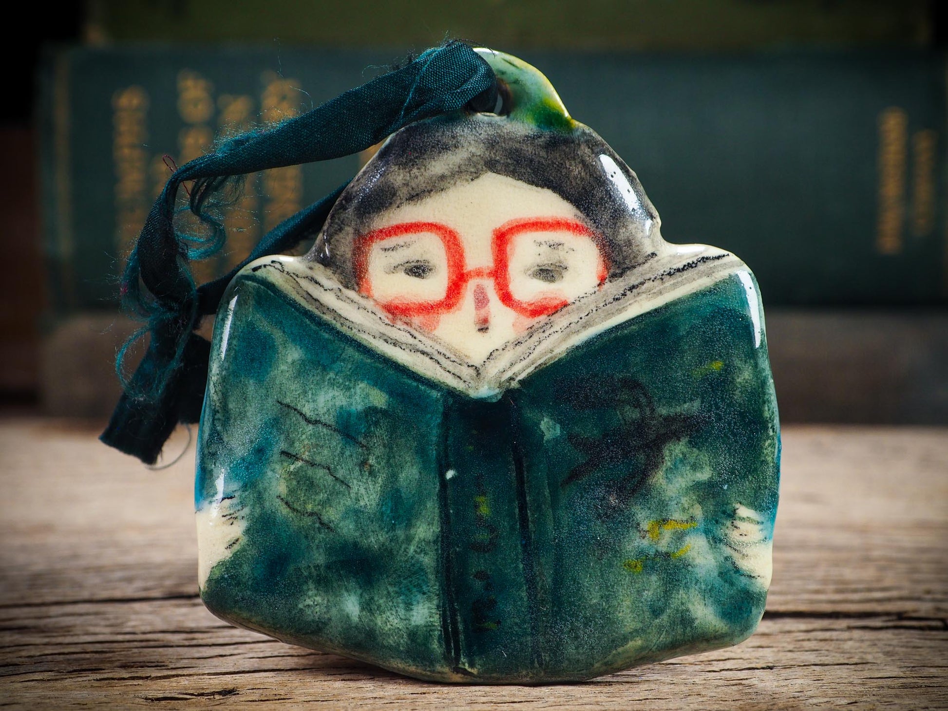 Handmade ceramic reader ornament with red glasses and green book, created by Idania Salcido aka Danita Art for Christmas decorating or gifting.