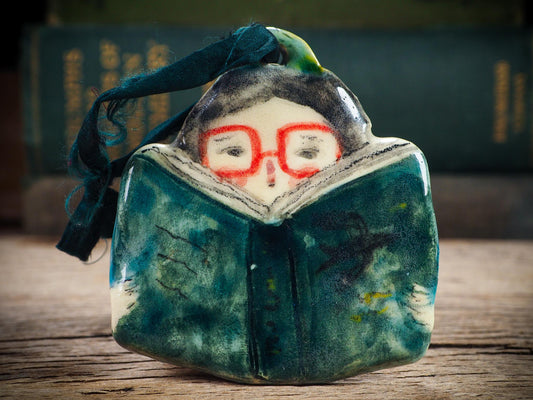 Handmade ceramic reader ornament with red glasses and green book, created by Idania Salcido aka Danita Art for Christmas decorating or gifting.