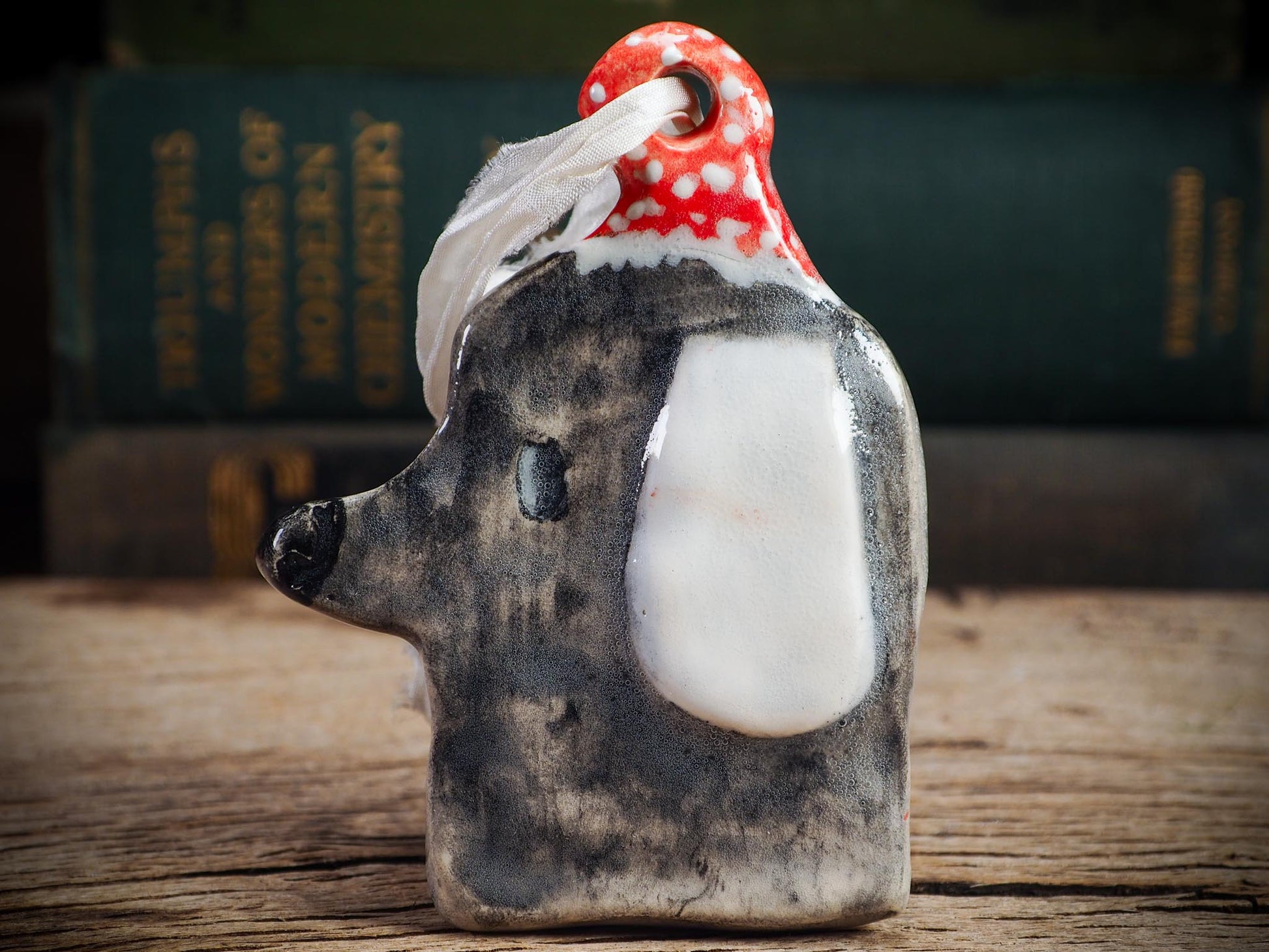 Handmade ceramic dog ornament with gray coat, white ear, and red dotted hood, created by Idania Salcido aka Danita Art for Christmas decorating or gifting.