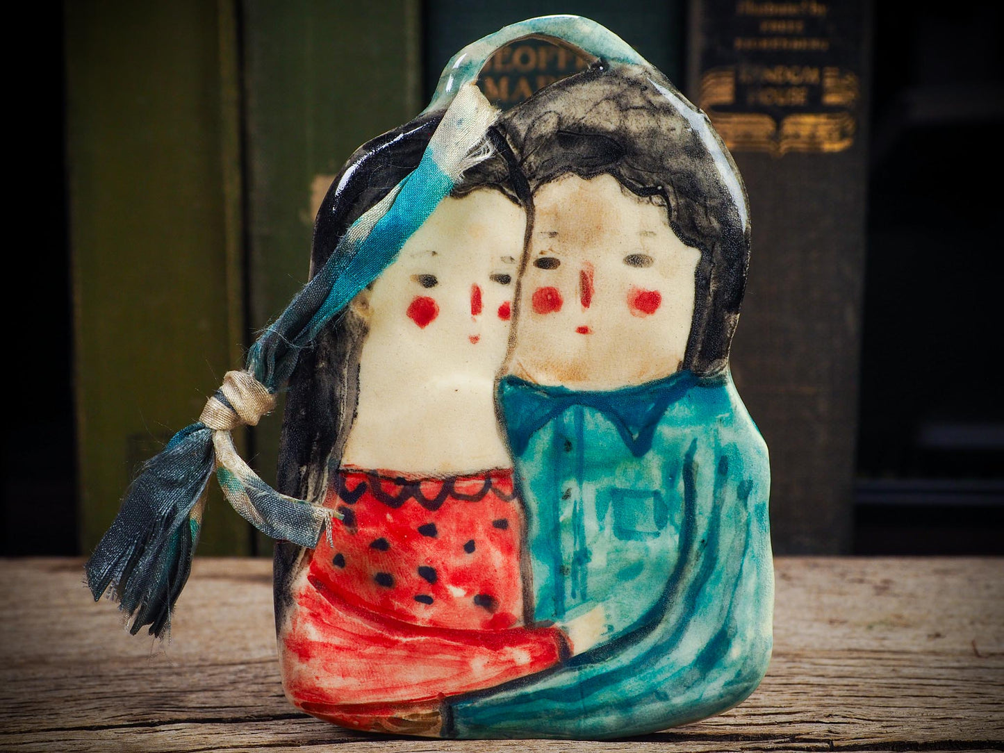 Handmade ceramic winter couple ornament with painted scarves and ribbon, created by Idania Salcido aka Danita Art for Christmas decoration or gifting.