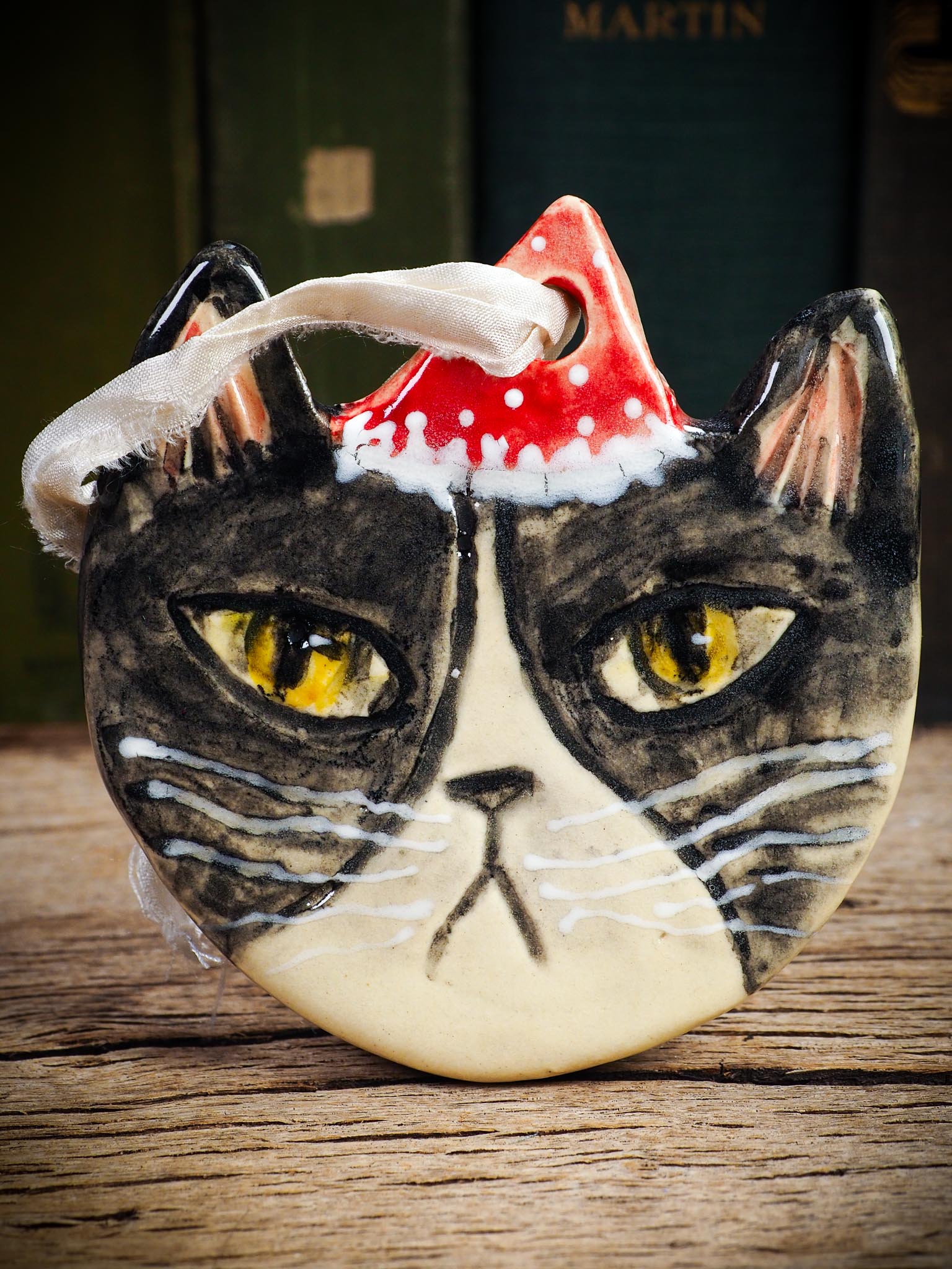 Handmade ceramic black and white tuxedo cat ornament with red holiday hat and ribbon, created by Idania Salcido Danita Art for Christmas decoration or gifting.
