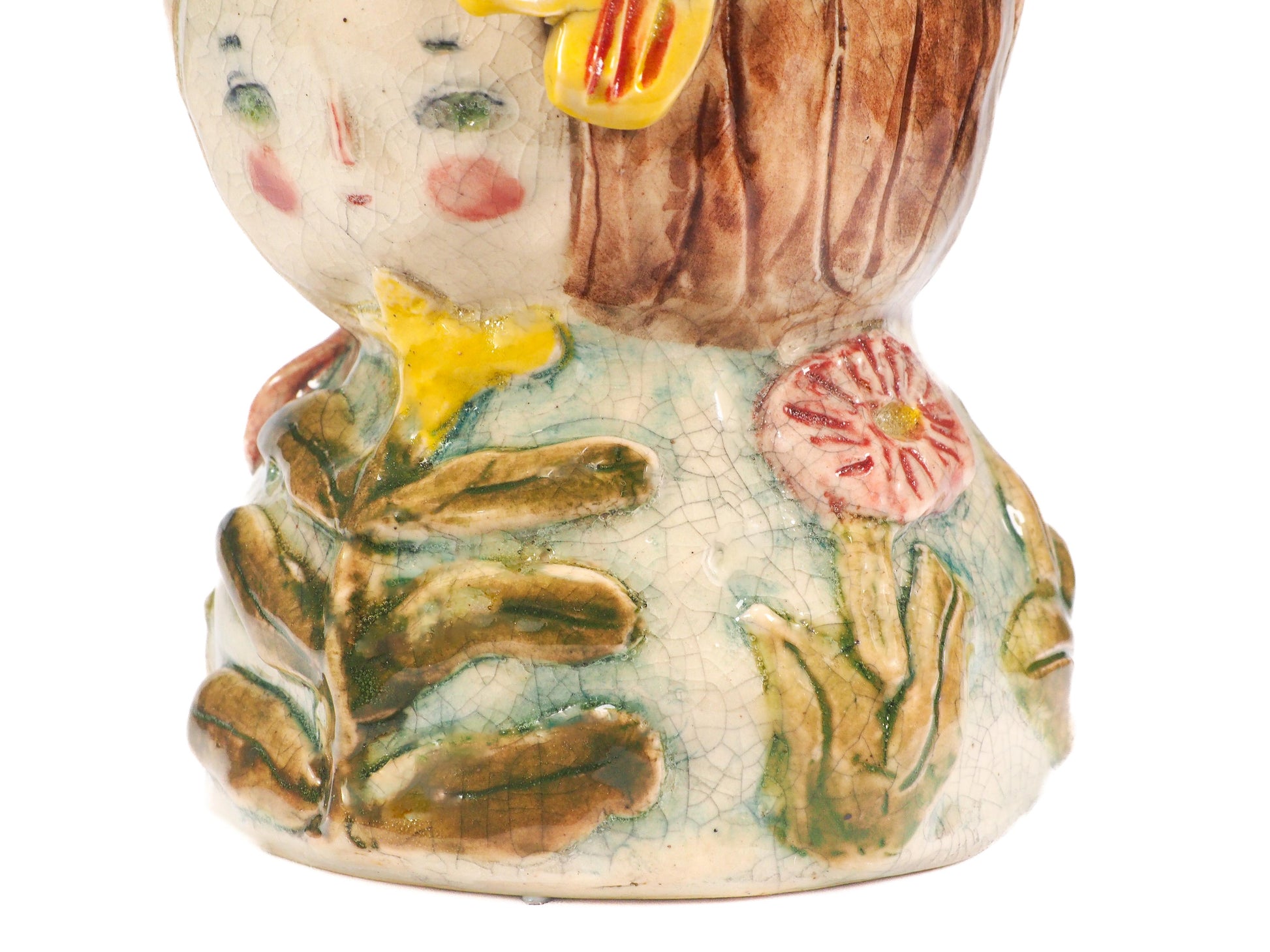A close-up of a hand-painted ceramic figurine featuring a stylized face with green eyes, pink cheeks, brown hair, a yellow flower, and a green leafy body with a pink flower by Idania Salcido (Danita Art).
