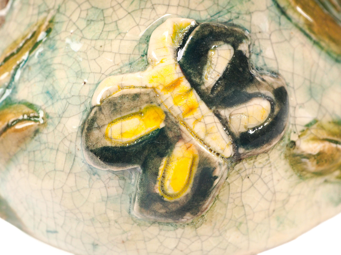 A close-up of a hand-painted ceramic figurine featuring a stylized face with green eyes, pink cheeks, brown hair, a yellow flower, and a green leafy body with a butterfly by Idania Salcido (Danita Art).