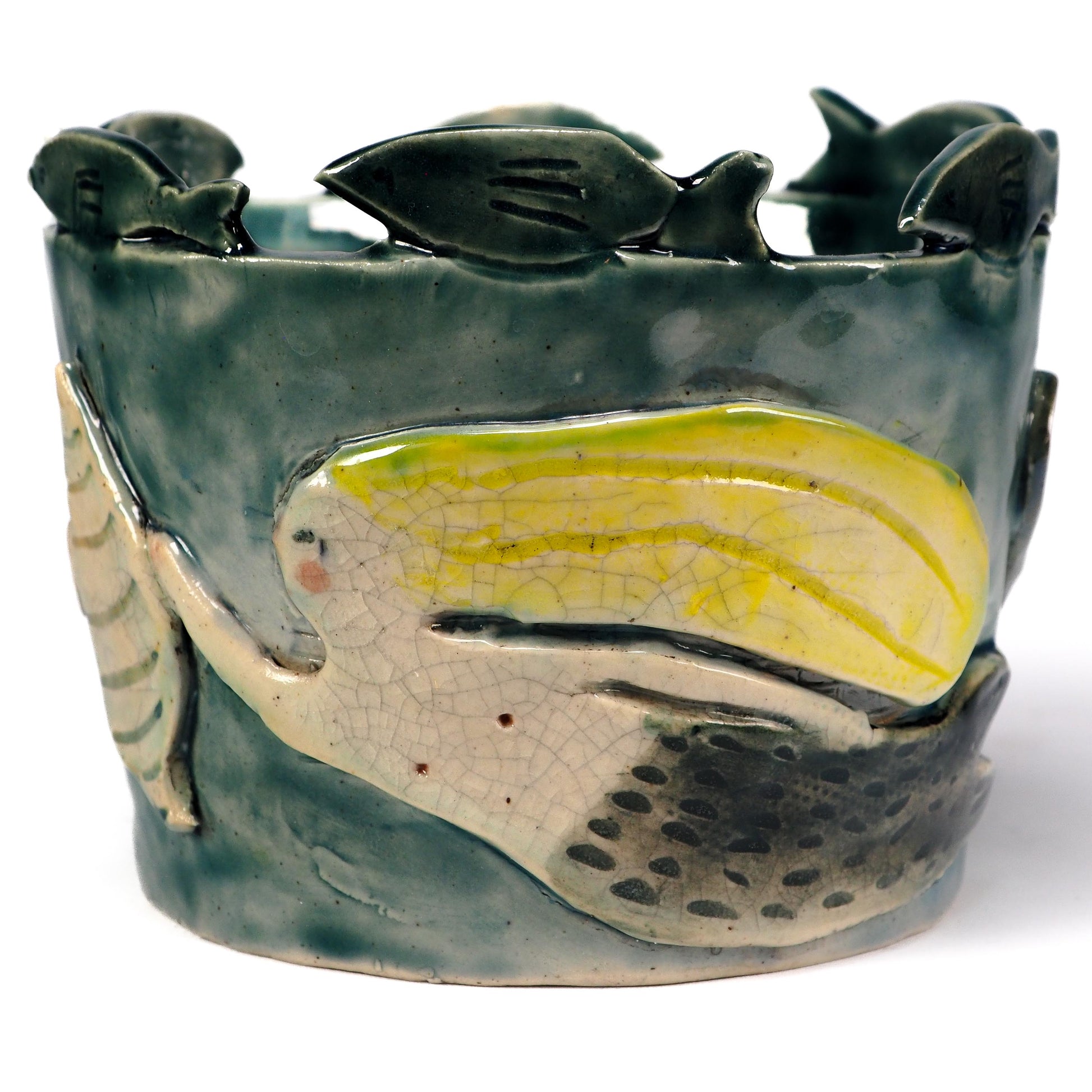 A handmade ceramic vase featuring a stylized fish design with a glossy yellow and cream body, set against a textured dark green glazed background with small fish figures along the top edge by Idania Salcido (Danita Art)