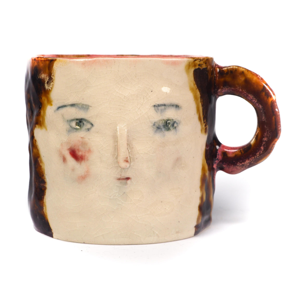 A handmade ceramic mug featuring a stylized face with green eyes, a painted red cheek, and a textured brown and cream glaze by Idania Salcido (Danita Art).