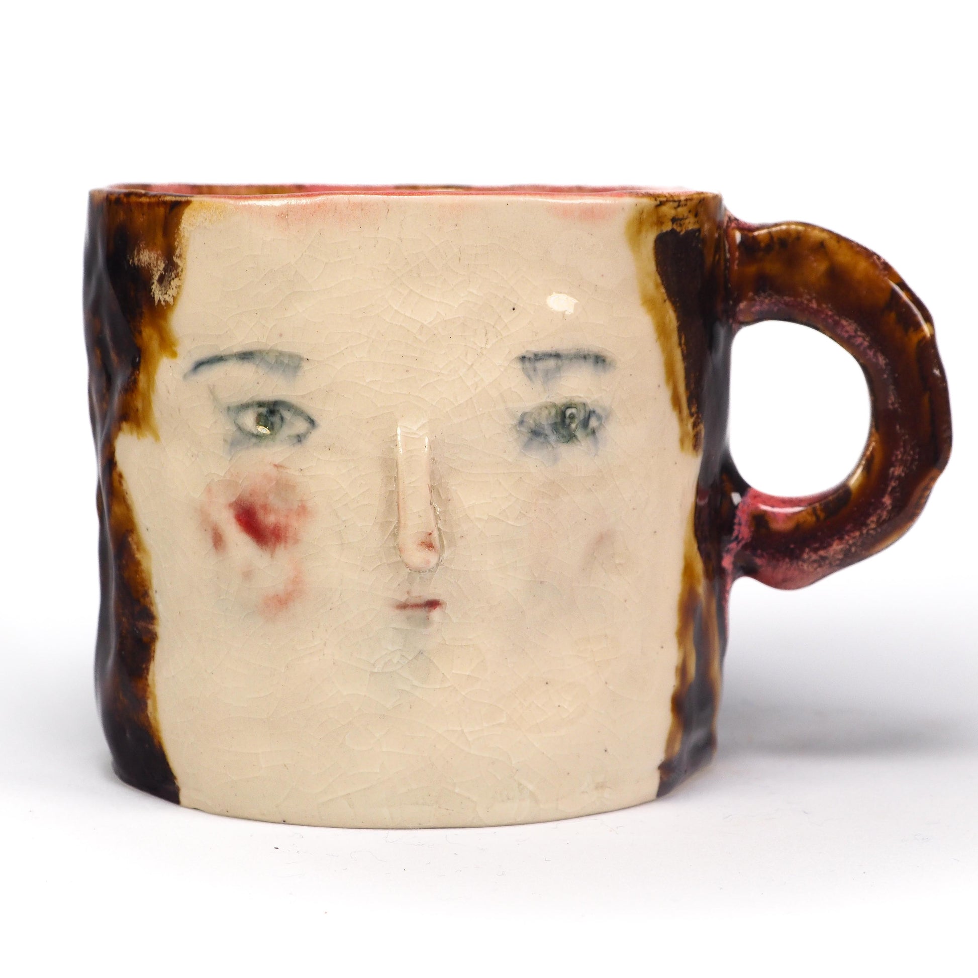 A handmade ceramic mug featuring a stylized face with green eyes, a painted red cheek, and a textured brown and cream glaze by Idania Salcido (Danita Art).