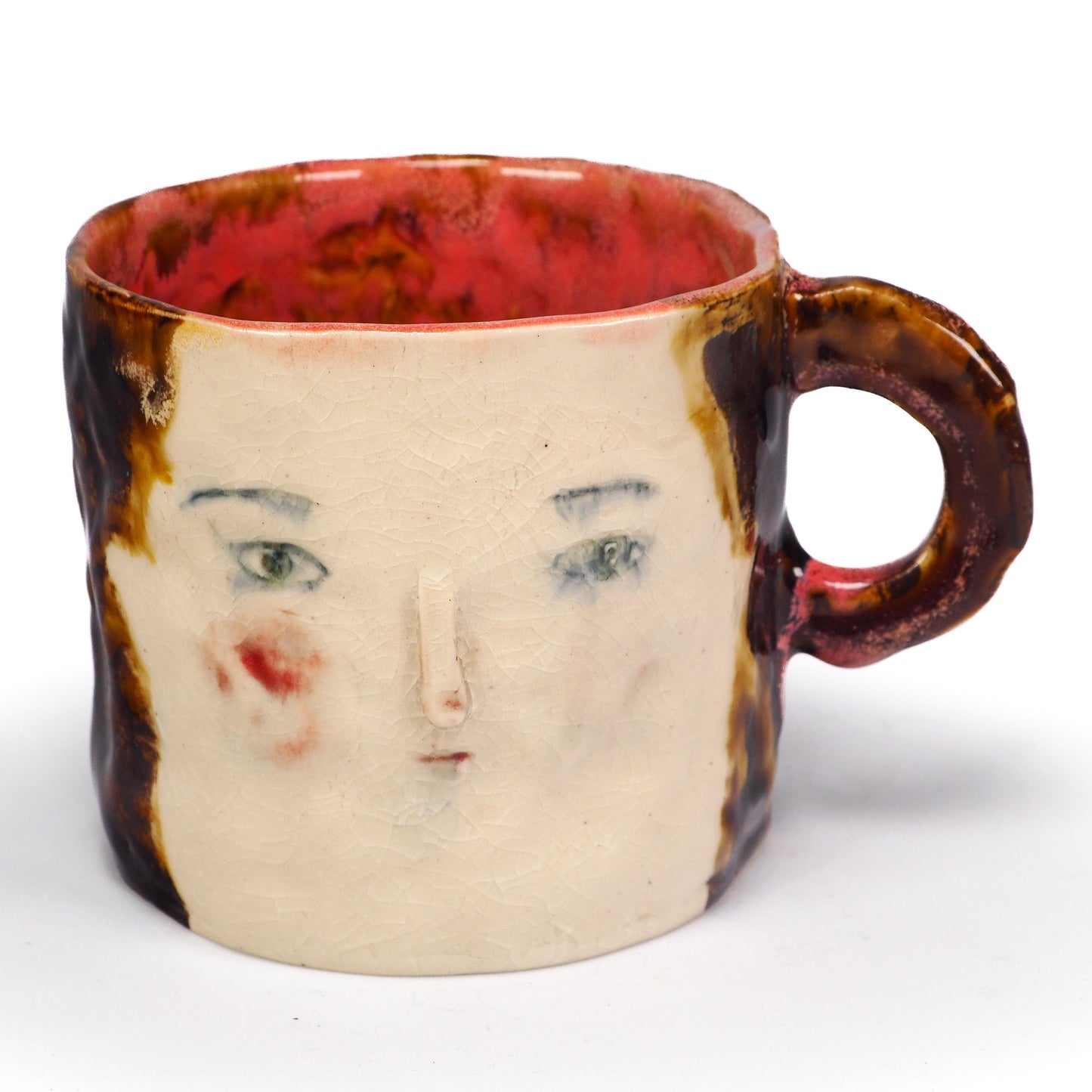 A handmade ceramic mug featuring a stylized face with green eyes, a painted red cheek, and a textured brown and cream glaze by Idania Salcido (Danita Art).