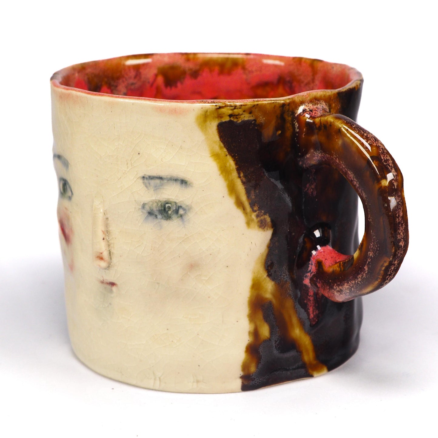 A handmade ceramic mug featuring a stylized face with green eyes, a painted red cheek, and a textured brown and cream glaze by Idania Salcido (Danita Art).
