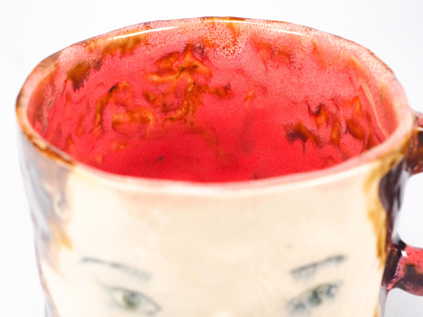 A handmade ceramic mug featuring a stylized face with green eyes, a painted red cheek, and a textured brown and cream glaze by Idania Salcido (Danita Art).