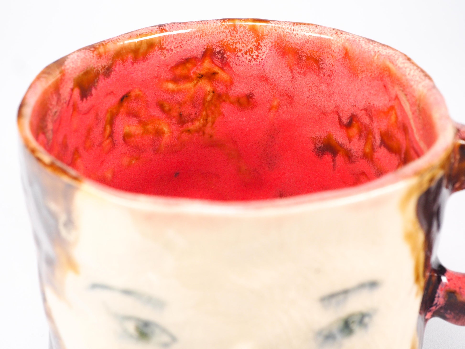 A handmade ceramic mug featuring a stylized face with green eyes, a painted red cheek, and a textured brown and cream glaze by Idania Salcido (Danita Art).