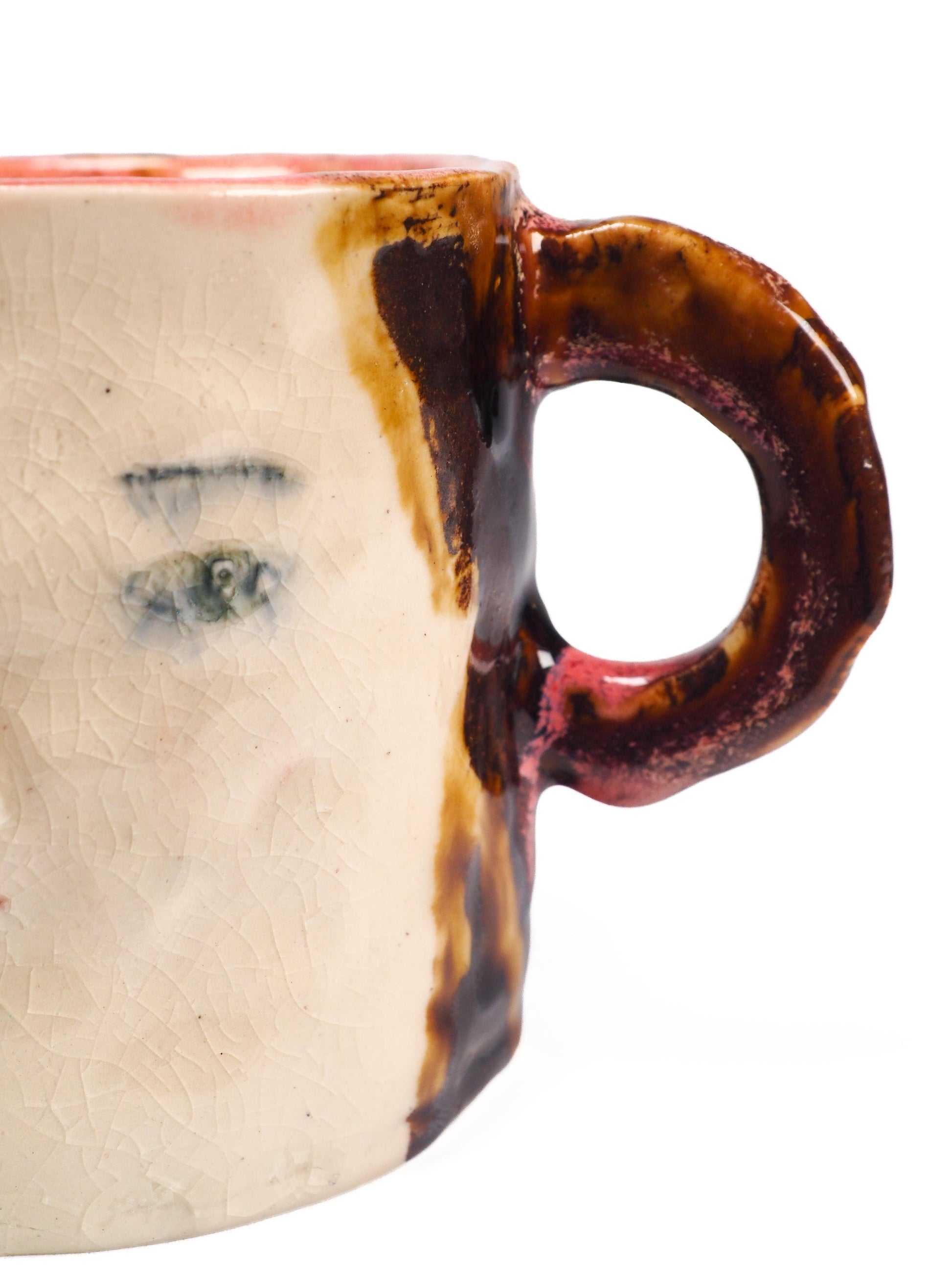 A handmade ceramic mug featuring a stylized face with green eyes, a painted red cheek, and a textured brown and cream glaze by Idania Salcido (Danita Art).