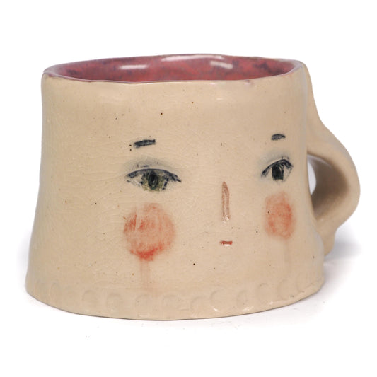 A handmade ceramic mug with a face design, featuring a beige stoneware body, hand-painted black eyes and eyebrows, pinkish-red cheeks, and a reddish-brown interior by Idania Salcido (Danita Art).