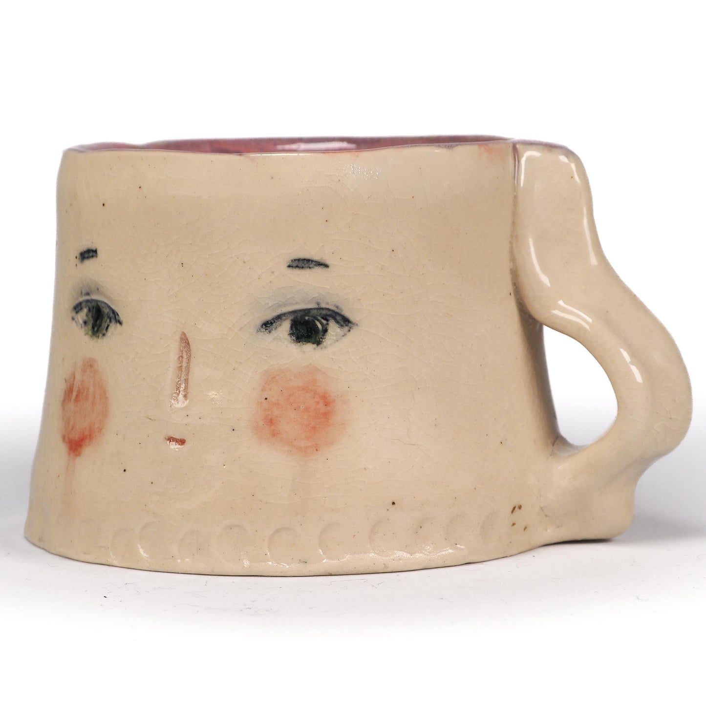 A handmade ceramic mug with a face design, featuring a beige stoneware body, hand-painted black eyes and eyebrows, pinkish-red cheeks, and a reddish-brown interior by Idania Salcido (Danita Art).