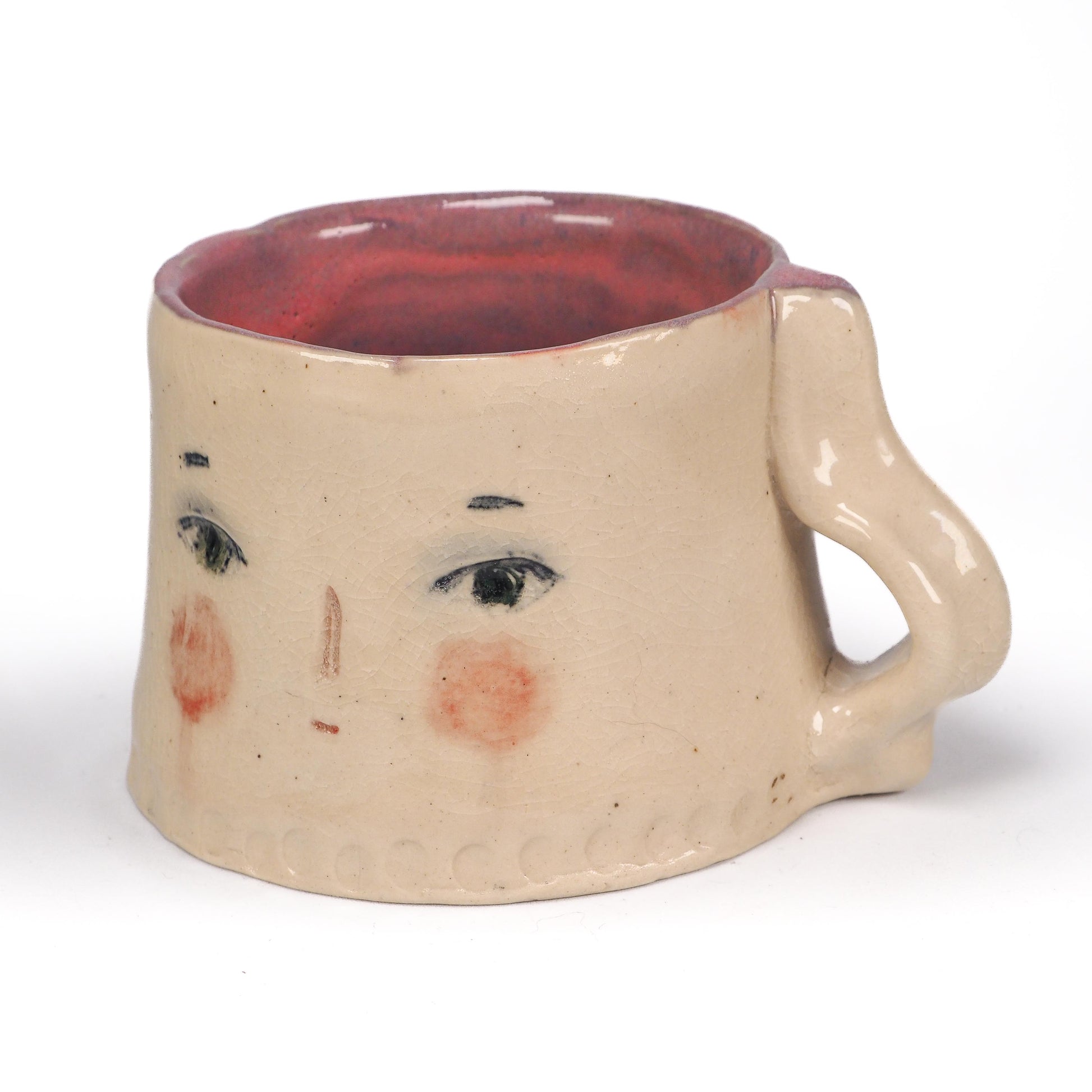 A handmade ceramic mug with a face design, featuring a beige stoneware body, hand-painted black eyes and eyebrows, pinkish-red cheeks, and a reddish-brown interior by Idania Salcido (Danita Art).