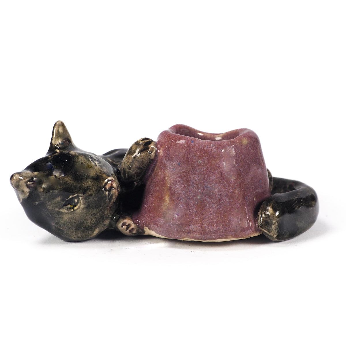A ceramic cat figurine lying down, holding a small purple bowl or cup in its paws. The cat's head and body are glazed in a dark metallic bronze-black, while the bowl has a textured, speckled purple glaze, by Idania Salcido (Danita Art).