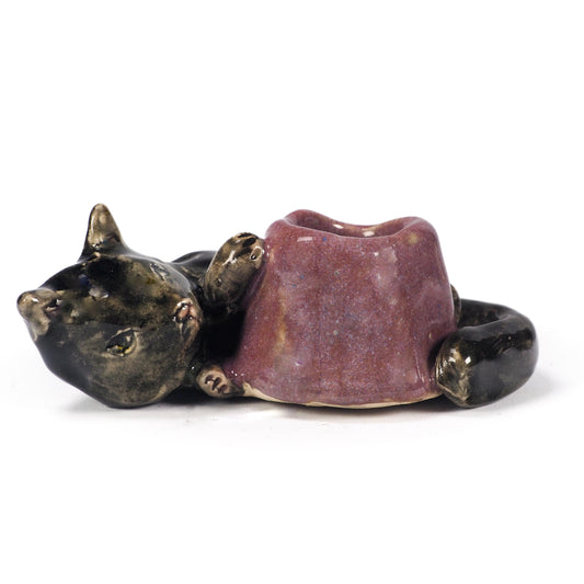 A ceramic cat figurine lying down, holding a small purple bowl or cup in its paws. The cat's head and body are glazed in a dark metallic bronze-black, while the bowl has a textured, speckled purple glaze, by Idania Salcido (Danita Art).